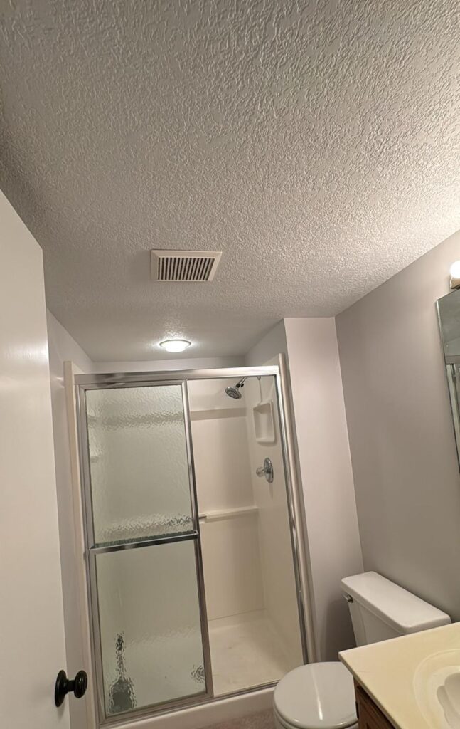Small bathroom with freshly painted light gray walls, a textured ceiling, and a glass sliding shower door next to a toilet and vanity.