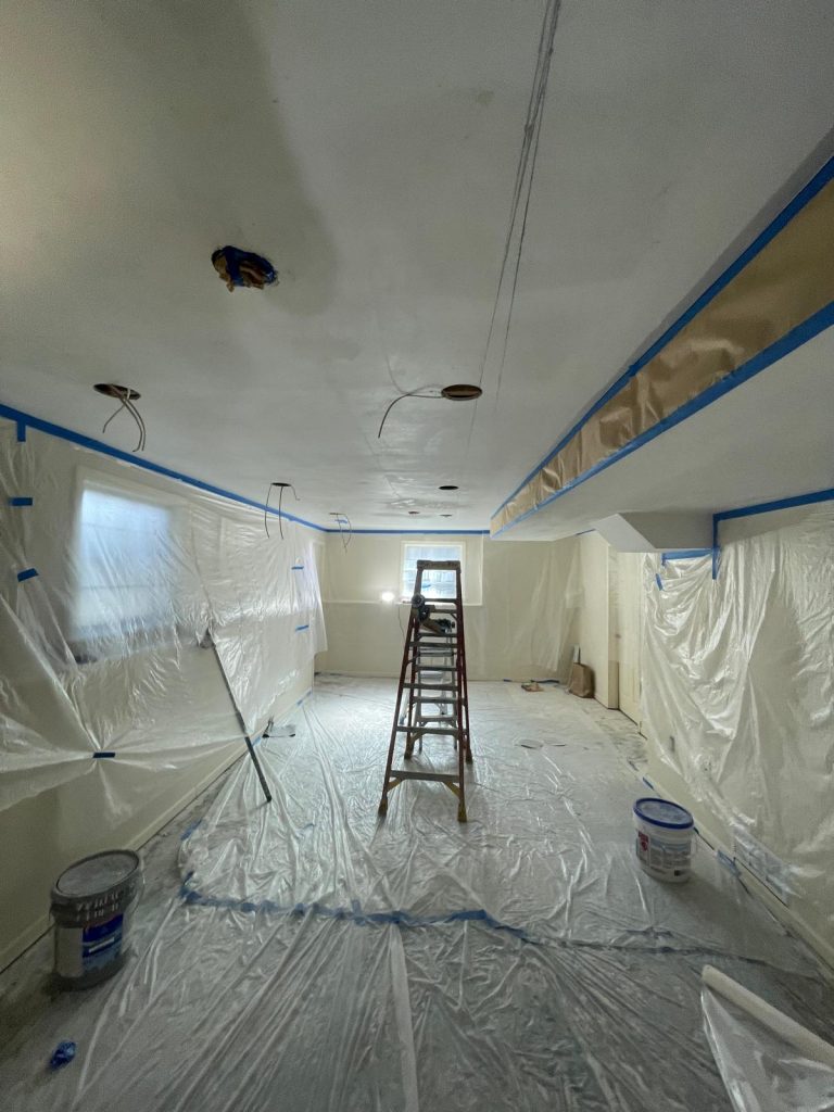 A room fully covered with plastic sheeting and painter’s tape while a ladder stands in the center, showing a ceiling painting project in progress with recessed lighting cutouts visible.